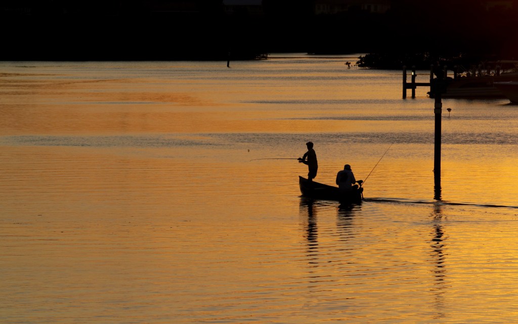 sunset photograph of two people on a row boat in a river silhouetted against the orange colored water reflecting the sky.